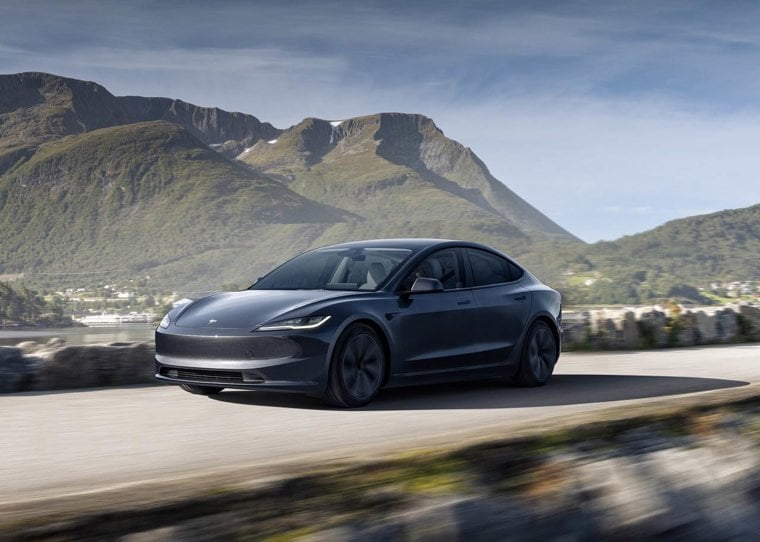 Grey Tesla Model 3 driving on a road with hills in the background