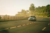 A modern white car drives along a scenic coastal road lined with palm trees under a clear sky