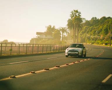 A modern white car drives along a scenic coastal road lined with palm trees under a clear sky
