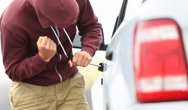 View down the side of a car to a man in a hooded top breaking into a car with a screwdriver in order to steal it.
