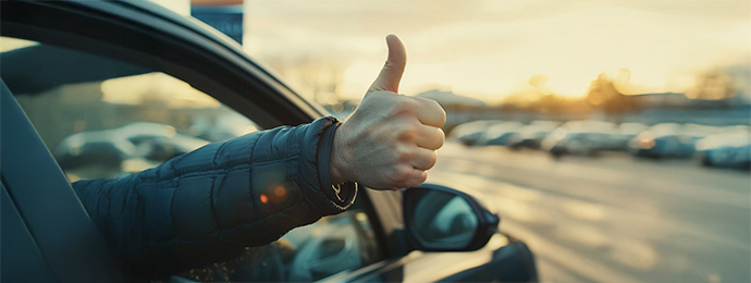 Close-up of a driver giving a thumbs-up out of a car window at a dealership car park during sunset
