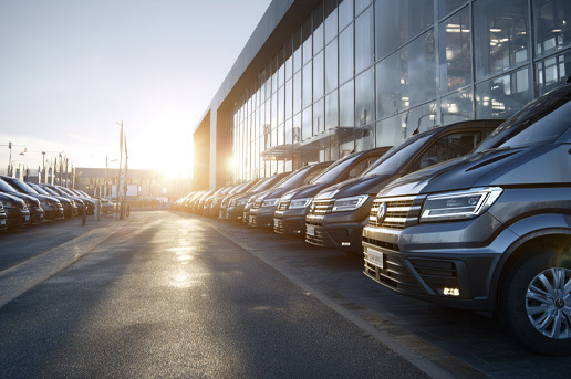 A wide angle view of a modern vehicle forecourt filled with rows of clean, new vans parked in neat lines outside a large glass fronted dealership