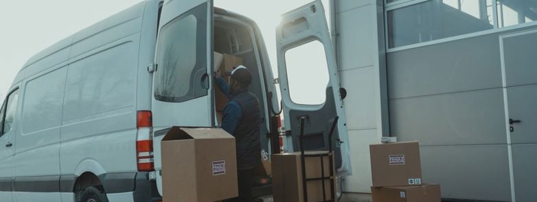 Courier loading cardboard boxes into a delivery van outside a warehouse.
