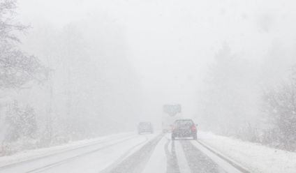 Heavy snowfall covers a road surrounded by a winter forest, with cars navigating the snowy conditions.