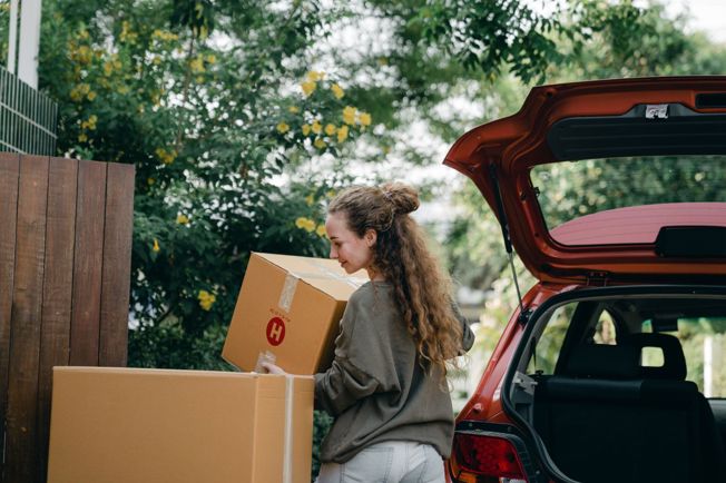 Happy young woman unloading cardboard boxes from car on a sunny day.