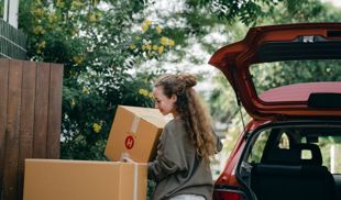 Happy young woman unloading cardboard boxes from car on a sunny day.