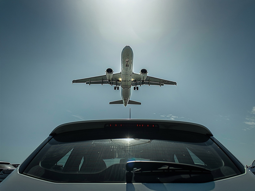 Plane flying low over car park, viewed from behind under bright sky