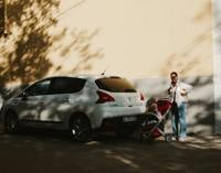 Mother with stroller and child beside a white car on a sunlit street
