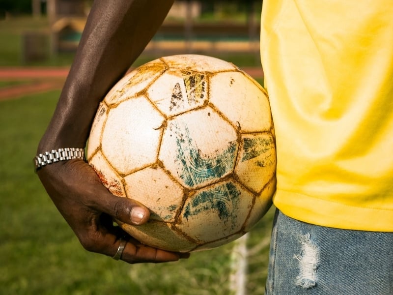 Close-up of a person holding a worn soccer ball on a sports field outdoors.