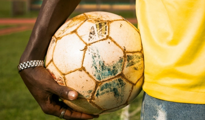 Close-up of a person holding a worn soccer ball on a sports field outdoors.