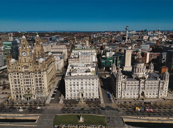 Aerial view of Liverpool featuring the Three Graces, buildings, roads, and skyline under a bright blue sky