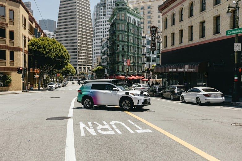 self-driving car driving down a street next to tall buildings