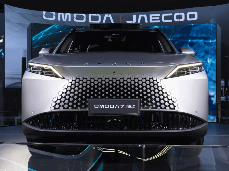 ront view of the Omoda 7 SUV, featuring a three-dimensional geometric grille, slim LED headlights, and metallic finish in an automotive show booth.