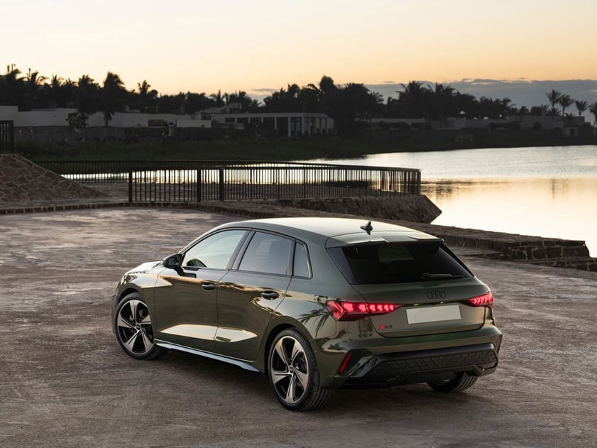 Rear view of a 2025 Audi A3 Sportback parked by a waterfront at sunset, showcasing modern hatchback design