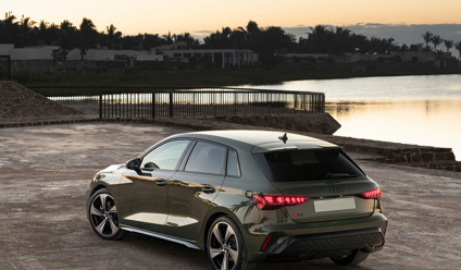 Rear view of a 2025 Audi A3 Sportback parked by a waterfront at sunset, showcasing modern hatchback design