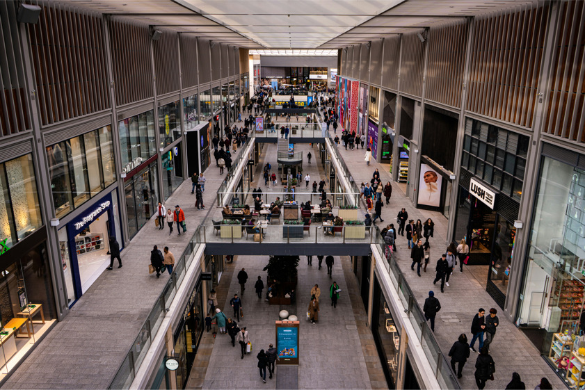 Busy UK shopping centre interior with two levels of retail stores, shoppers walking and sitting in communal seating areas, showing modern retail experience