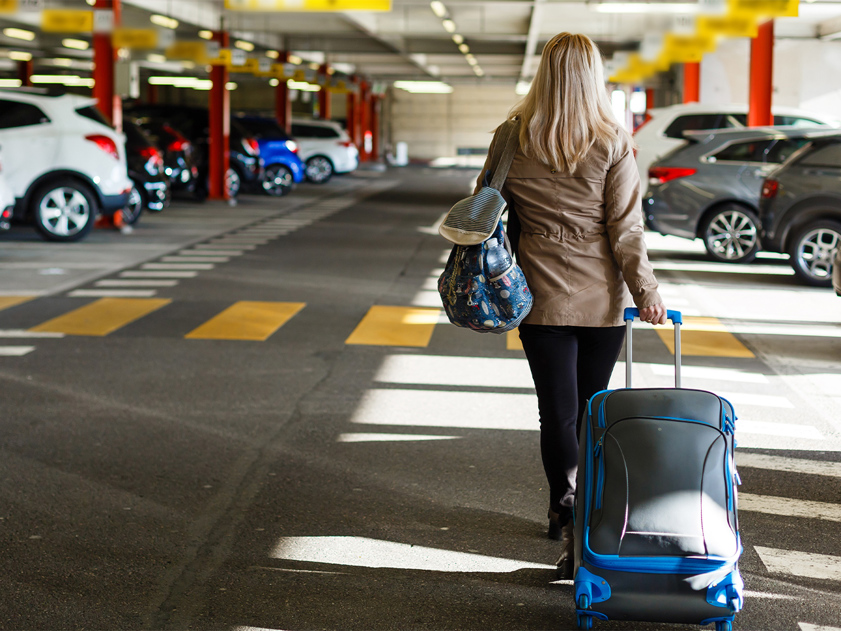 Woman walking through airport car park with suitcase, heading towards terminal