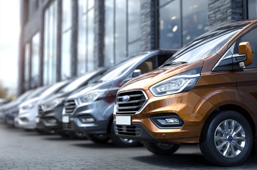 A photorealistic image of a row of generic new vans parked at a UK-style dealership, blurred with large glass windows and a grey brick exterior.