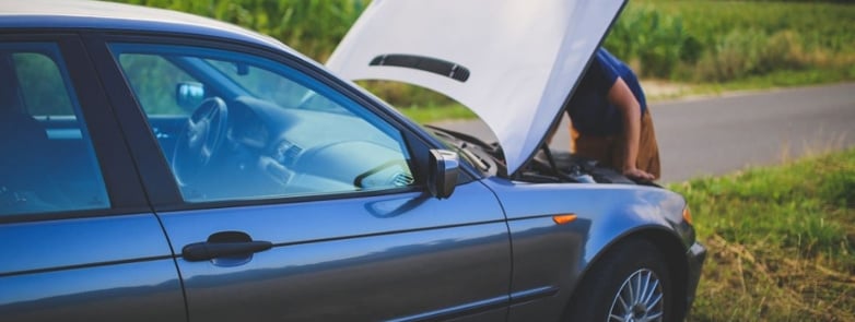 Man checking car engine with hood open by the side of a rural road.