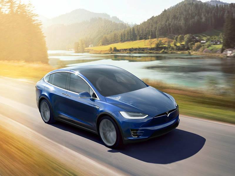 blue tesla model x driving on the road with lake behind