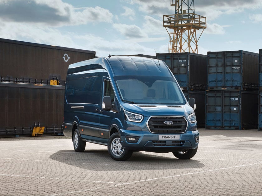 Front-side view of a blue Ford Transit parked in a shipyard with shipping crates in the background