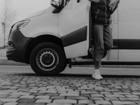 Monochrome image of a person stepping out of a van onto cobblestone pavement.