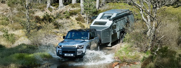 Land Rover Defender 110 towing an off-road caravan through a forest trail and shallow water crossing