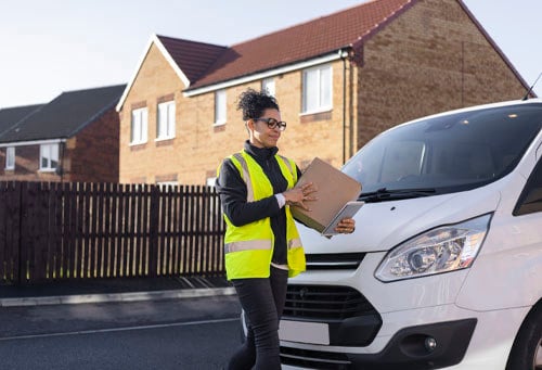 female postal worker delivering a parcel to a residential address in an urban area She is walking around the from of the delivery van past the camera.