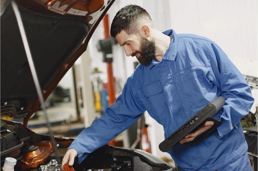 Mechanic in blue overalls looking an an engine bay with a tablet in his hand