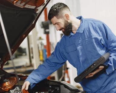 Mechanic in blue overalls looking an an engine bay with a tablet in his hand
