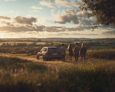 Group of young adults standing next to an SUV on a grassy hill at sunset, overlooking rolling countryside and fields