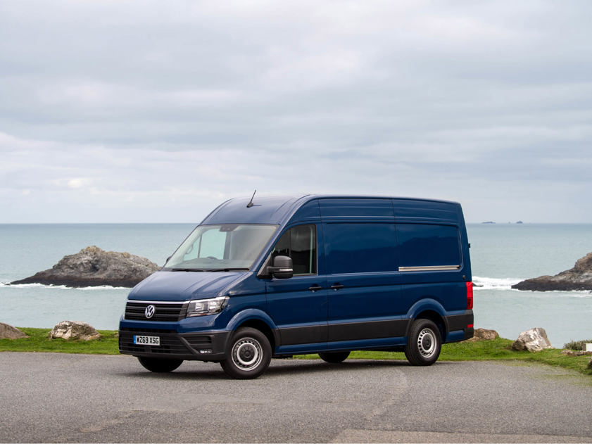 Front-side view of a blue Volkswagen Crafter parked by the coast with the sea and rocks in the background